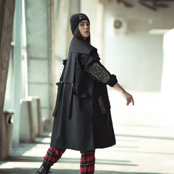 Une femme portant un long manteau noir, un pantalon à carreaux et un bonnet regarde en arrière tout en marchant dans un espace intérieur de style industriel.