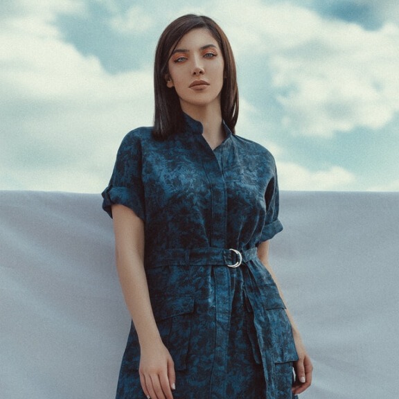 Femme portant un bonnet sarcelle avec des écussons en forme de nuages portant l'inscription "OKAY ? OKAY" et une veste orange, regardant la caméra par-dessus son épaule.