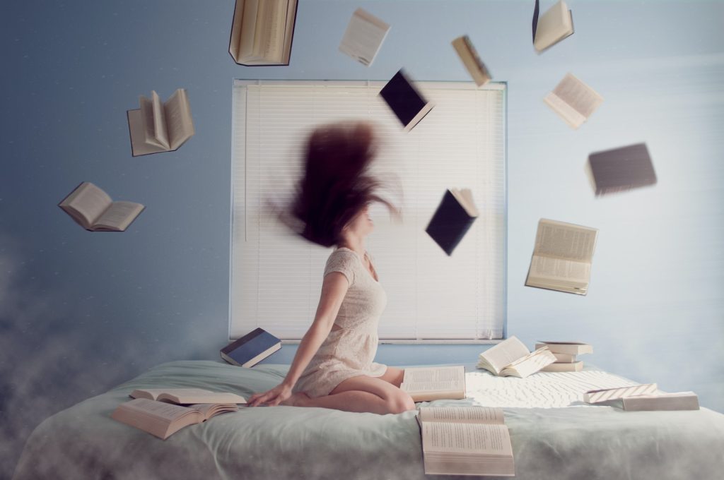 Une fille assise sur un lit avec des livres volant autour d’elle.